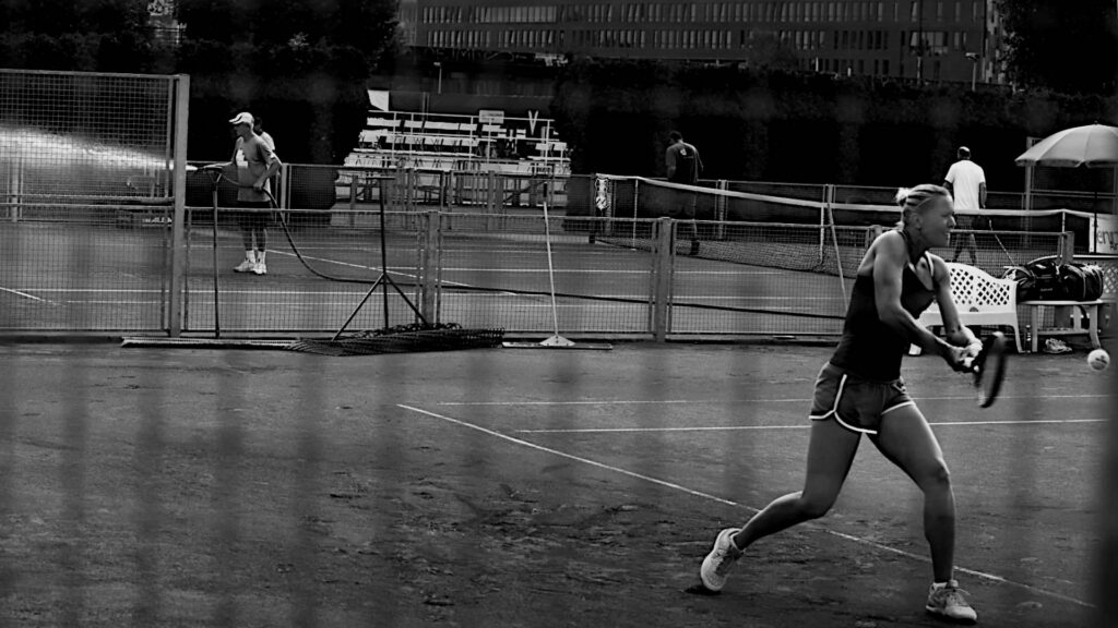 Prague 2017 - Being deliberately photographed through a grid, Lucie Hradecka giving it her all on the front court while the next is being prepared for an upcoming metch. In the background one can see trees, in the center the stands of another court, almost in the dark a bridge over river Moldau and a big office building on the other bank. This Štvanice island venue, where the folks of the I. Český lawn-tennis klub (I. ČLTK Praha) hospitably granted me accreditations twice, has been one of my favorite 'workplaces'.